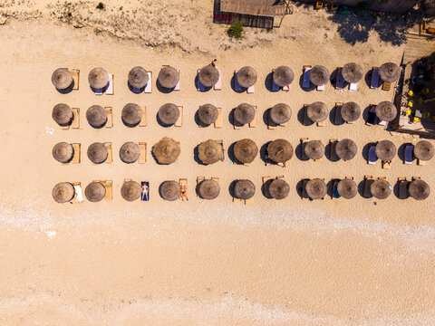 Aerial view of rows of straw umbrellas and sun loungers arranged on the sandy beach with people relaxing under the sun in Sarande, Vlore County, Albania.