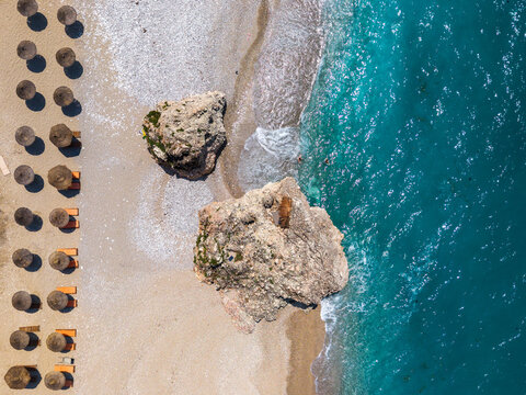 Aerial view of Sarande beach with rows of straw umbrellas and sun loungers on the sand, large rock formations near the turquoise shoreline in Sarande, Vlore County, Albania.