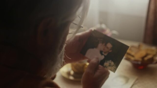 Over shoulder of senior Caucasian man sitting alone at table, holding and gently touching old wedding photograph. Ideal for senior care services, grief counseling, or retirement planning campaigns