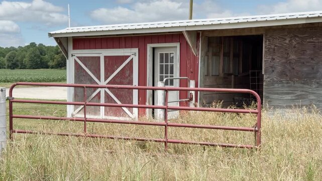 Small red horse barn run-in shed with white cross beams stands behind rusty gate with pasture in foreground