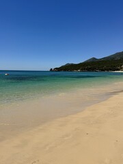 Obraz premium Praia de areia dourada banhada por águas cristalinas, sob um céu azul sem nuvens. Ambiente sereno e convidativo, ideal para representar férias, verão e relaxamento.
