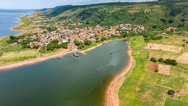 Aerial view of a lakeside village with small houses, a ferry landing, and agricultural fields along the shoreline of Lake Volta in Kwahu Afram Plains South, Eastern Region, Ghana.