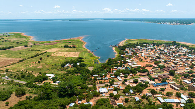 Aerial view of a town situated along the shores of a large lake with surrounding green fields, scattered trees, and a road network, Kwahu Afram Plains South, Eastern Region, Ghana.