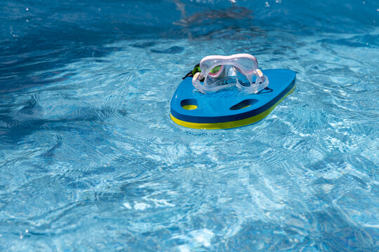 Swimming goggles are resting on a floating kickboard on the surface of a swimming pool. The calm, clear water creates a relaxed summer atmosphere, highlighting poolside leisure and swim training equip