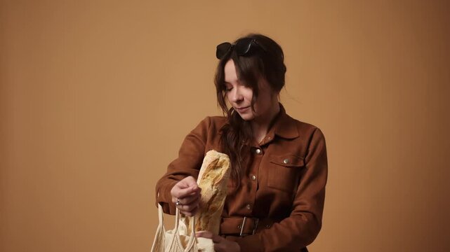 Stylish woman putting a fresh baguette and groceries into a reusable net bag for sustainable shopping against a brown background