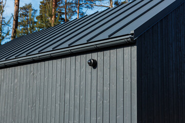 Charred timber wall with vertical cladding supports a matte metal roof and gutter. A dome camera sits on the eave. Conifer trunks and daylight suggest a wooded setting. © True Pixel Art