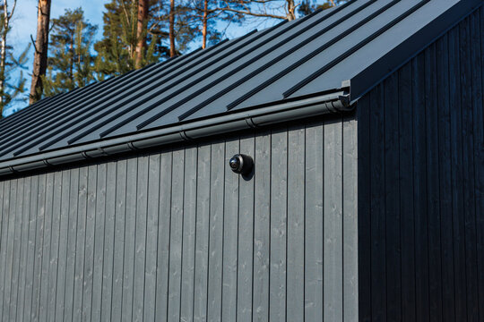 Charred timber wall with vertical cladding supports a matte metal roof and gutter. A dome camera sits on the eave. Conifer trunks and daylight suggest a wooded setting.