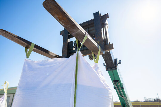 A green telehandler lifts a large white bulk bag on weathered tines. Low angle view shows taut straps, sunlit metal, and midday light in an outdoor storage yard.