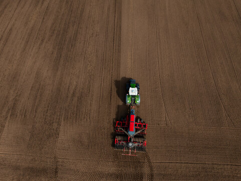 Aerial view shows a green tractor towing a red cultivator making parallel furrows across a vast brown field, late afternoon light casts long shadows and clear tire tracks.