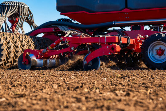 Red machinery pulls a disc harrow across dry soil as clods scatter. Large treaded tires, hydraulic linkages, staggered discs, and seed tubes show modern field work in daylight.