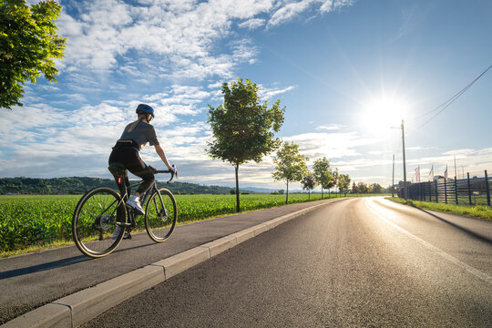 Female cyclist with road bike riding on asphalt path at sunset in summer in Slovenia. Rear view of woman cycling in helmet near green field, active sport lifestyle, bright evening sun sky landscape
