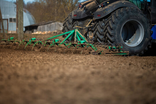 A powerful tractor with oversized rear tires pulls a green multi row cultivator, kicking up clods in brown soil near outbuildings, in warm daylight, low angle view.