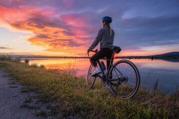 Female cyclist with road bike at lake side at sunset in summer. Slovenia. Rear view of woman riding bicycle in helmet on gravel path near water, active sport lifestyle, colorful twilight sky landscape