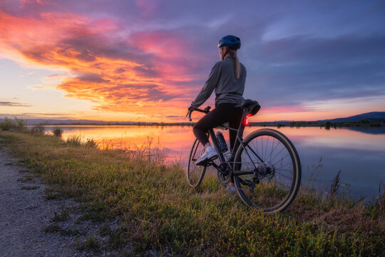 Female cyclist with road bike at lake side at sunset in summer. Slovenia. Rear view of woman riding bicycle in helmet on gravel path near water, active sport lifestyle, colorful twilight sky landscape