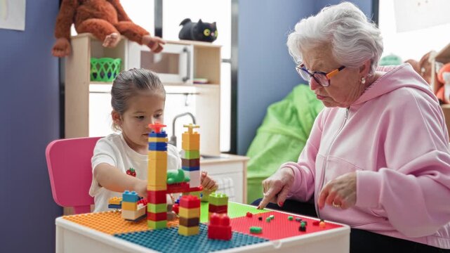 Elderly woman and little girl stack colorful building blocks and share a toy handed across the table in playroom; tenderness bonding.