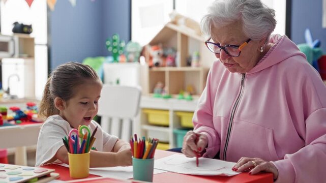 Elderly woman guides child's drawing with colored pencils and paper in a daycare building; intergenerational learning warmth.