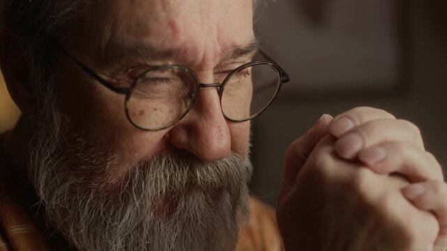 Closeup of senior Caucasian man praying with clasped hands, taking off glasses and rubbing forehead. Video ideal for mental health awareness, senior loneliness, or retirement support campaigns