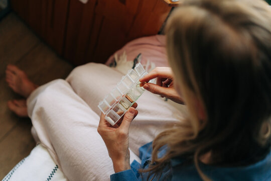 An unrecognizable woman holds a pillbox with pills in her hands while sitting in a room.