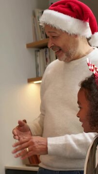 Vertical video: Clapping senior in holiday headwear offering cookie in kitchen, child leaning