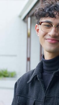 Vertical video: Camera filming man with glasses turning and laughing near building in jacket