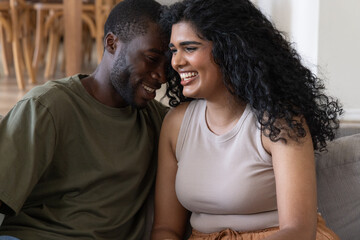 Obraz premium Diverse couple laughing and leaning together on light sofa in living room, wooden table behind