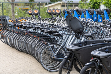 Fototapeta premium Row of parked bicycles in a bike-sharing station with various models and colors, surrounded by greenery and urban infrastructure in a city setting