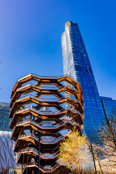 New York, United States - April 11, 2026: The golden honeycomb structure of the Vessel stands prominently in Hudson Yards, contrasted against the soaring blue glass facade of nearby skyscrapers.
