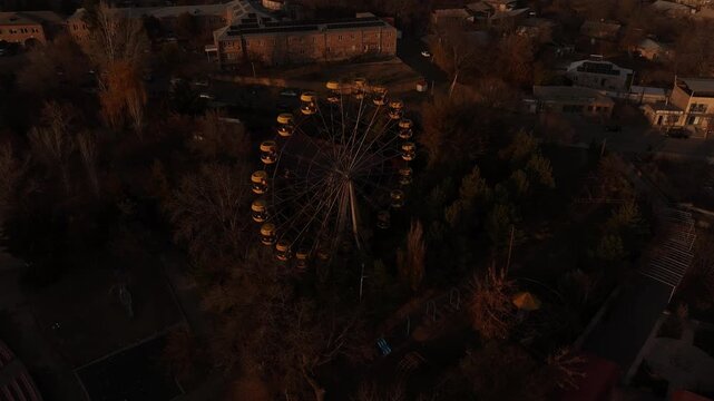 An aerial view of an old Ferris wheel with yellow cabins in an autumn park, gradually tilting up to reveal the scenic city surrounded by misty mountain landscape.

