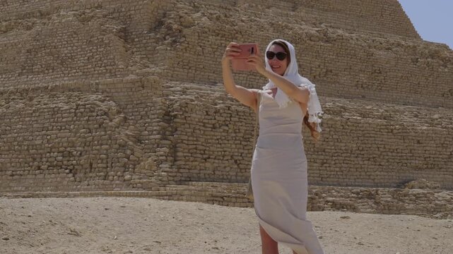 Slow motion 4K travel video of a female tourist taking a selfie with a smartphone in front of an ancient pyramid in Egypt, capturing a memorable vacation moment at a famous historical landmark.