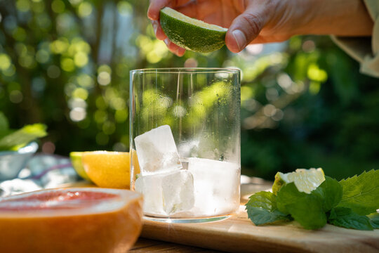 Person squeezing a fresh lime wedge over a glass with ice cubes to prepare a summer cocktail
