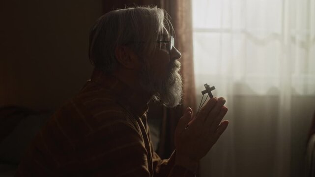 Side view of senior Caucasian man with beard and glasses praying while holding small wooden cross in clasped hands. Footage suitable for religious programs or spiritual wellness advertisements