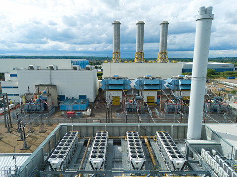 Aerial view of Rye House Power Station with its three tall chimneys, industrial cooling units, and electrical infrastructure under a cloudy sky in Hoddesdon, England, United Kingdom.