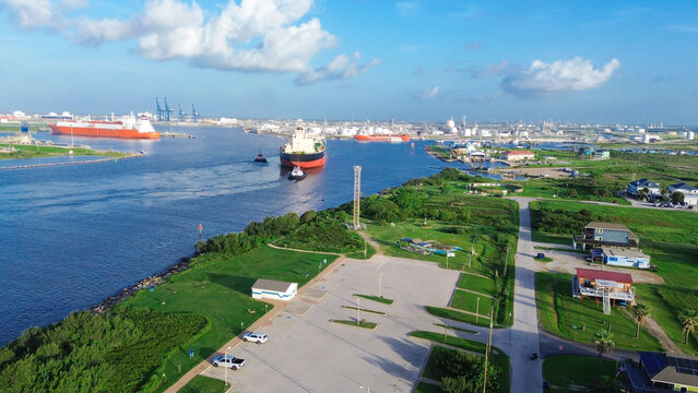 Massive marine vessels move toward industrial refineries as they pass through Harbor shipping lanes. Verdant shoreline vegetation contrasts with harbor infrastructure, Freeport, TX