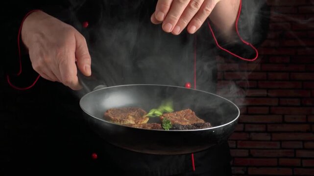 Chef in black uniform sprinkles fresh herbs onto sizzling meat in a frying pan, steam rising in a rustic kitchen setting with brick wall background