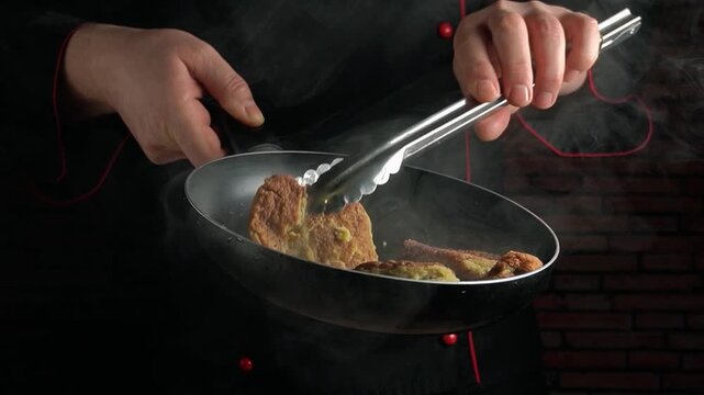 Chef in black uniform flips food in frying pan with tongs, steam rising, showcasing culinary skills in a professional kitchen setting