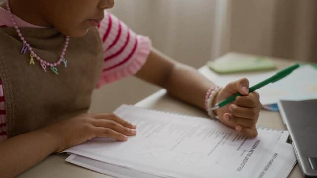 African American girl writing on math homework sheet with green pen at desk. Footage ideal for education marketing, e-learning platforms, and tutoring service advertisements