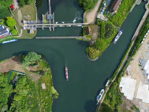 Aerial view of Feildes Weir Lock on the River Lee, featuring a narrowboat navigating the waterway, surrounding lush green trees, and calm water in Hoddesdon, England, United Kingdom.