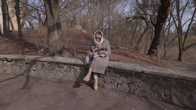 Inspired mature woman poet in coat with scarf writes poems sitting on parapet in spring park. Senior female author works on future novel plan in urban garden. Thoughts notices in diary