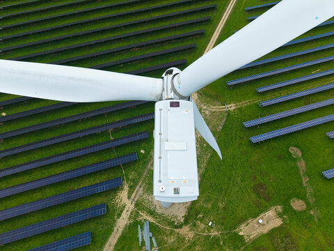 Aerial view of a wind turbine integrated within a solar farm featuring rows of photovoltaic panels on green grass in Chelveston, England, United Kingdom.