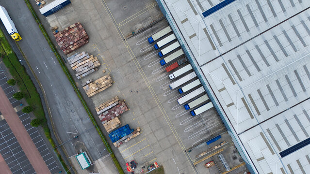 Aerial view of Kettering industrial warehouses featuring loading docks with parked semi-trucks, stacks of wooden pallets, and a large warehouse roof in Kettering, England, United Kingdom.