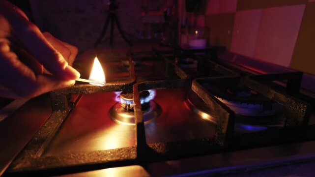 Hand uses a lit match to ignite the blue flame of a gas stove burner. Close-up shot captures the ignition process in a dark, atmospheric kitchen.