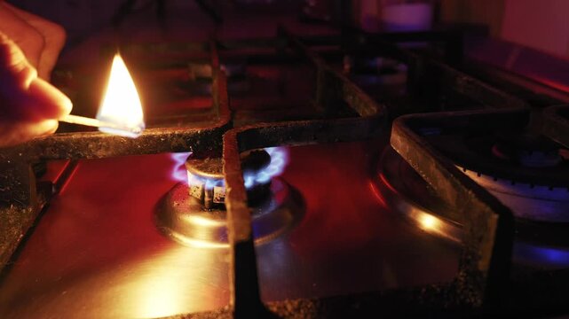 Hand uses a lit match to ignite two gas stove burners in sequence. Dramatic red and blue light illuminates the blue flames against the dark background.