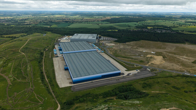 Aerial view of Corby Gate Business Park featuring large industrial warehouse facilities with expansive parking lots, surrounded by green fields in Corby, England, United Kingdom.