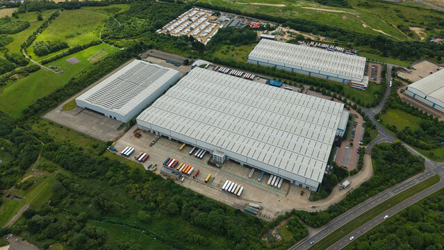 Aerial view of Corby Gate Business Park featuring large industrial warehouses, loading docks with parked trucks, and surrounding green fields in Corby, England, United Kingdom.