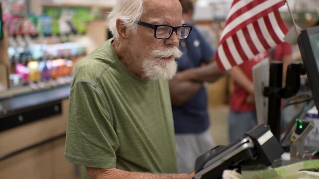 Elderly man stands at checkout counter in grocery store happily paying for his groceries with a credit card surrounded by shoppers