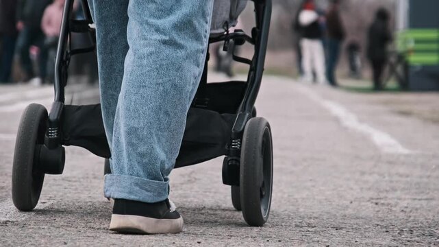 A low-section shot captures a woman's legs as she pushes a baby stroller on a city street. The blurred background draws attention to the motion of walking and the wheels.