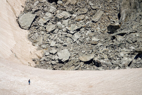 Aerial view of a lone hiker walking across a vast snowfield near a massive rocky moraine and glacier in Courmayeur, Aosta Valley, Italy.