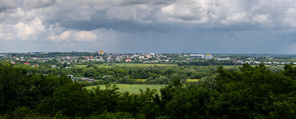 Obraz premium Pidvolochysk, Ternopil Oblast, Ukraine: A panoramic view of the town under dramatic storm clouds, a prominent golden-domed church and modern grain silos amidst vast green landscapes and fields