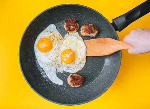 A child's hand flips fried eggs in a frying pan next to three cutlets on a yellow background. Breakfast, lunch, and dinner concept. Top view.