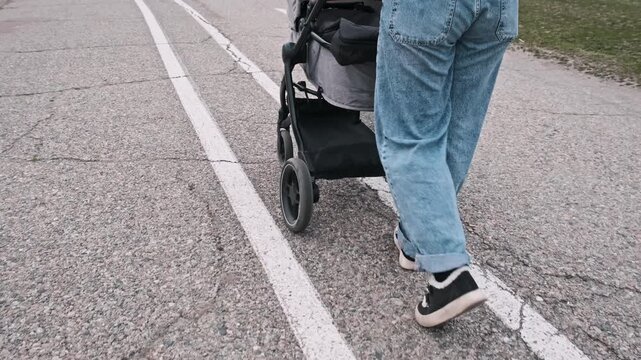 A woman in jeans and sneakers pushes a baby stroller along a marked asphalt road. The steady movement captures the casual pace of a daily walk in the city environment.